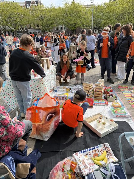 A lively outdoor party with people gathered in a public square surrounded by trees, with tables of food and gifts